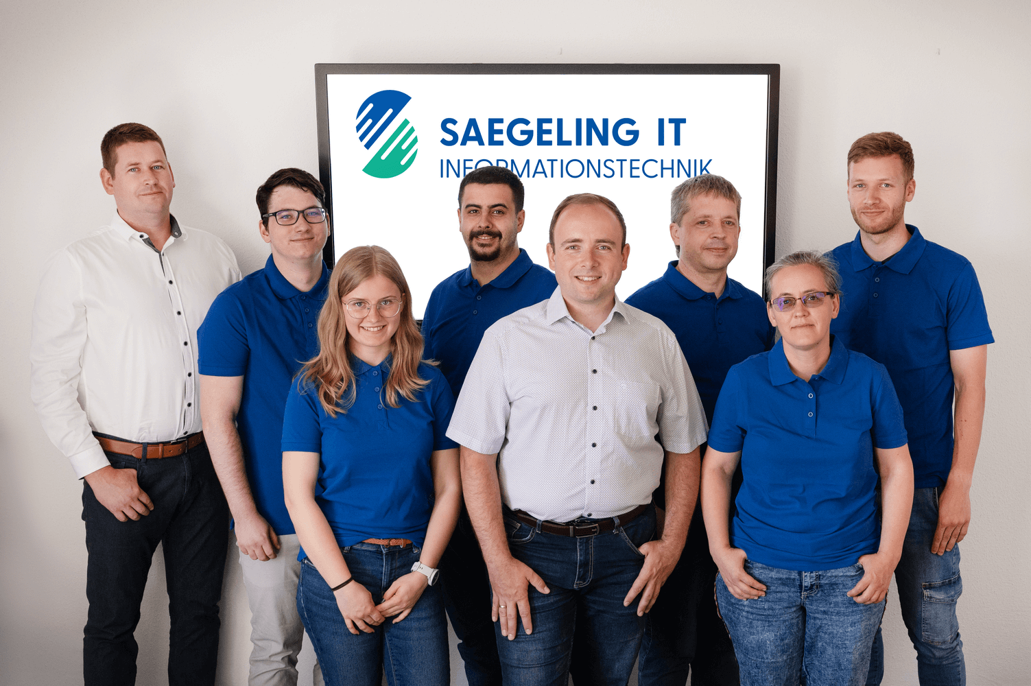 Das Bild zeigt ein achtköpfiges Team der Saegeling IT Informationstechnik vor einem Bildschirm mit Firmenlogo. Sechs Mitglieder tragen blaue Poloshirts, zwei heben sich mit hellen Hemden ab. Die Gruppe wirkt gemischt, freundlich und professionell. Das Bild zeigt ein achtköpfiges Team der Saegeling IT Informationstechnik vor einem Bildschirm mit Firmenlogo. Sechs Mitglieder tragen blaue Poloshirts, zwei heben sich mit hellen Hemden ab. Die Gruppe wirkt gemischt, freundlich und professionell.
